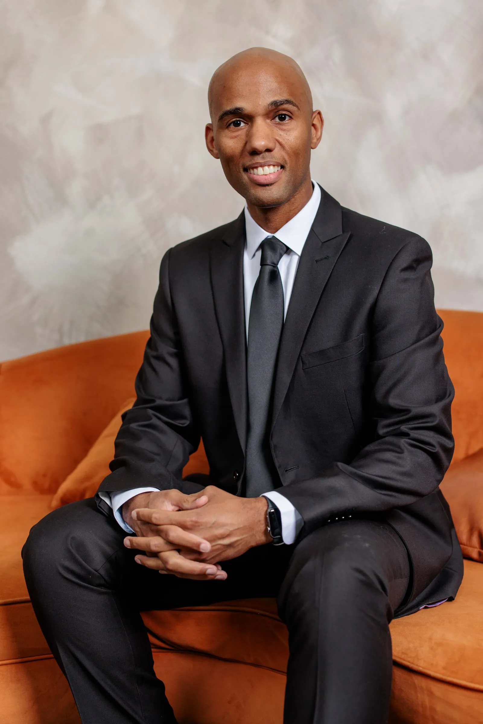 A man in a black suit and tie seated with hands clasped on an orange sofa.