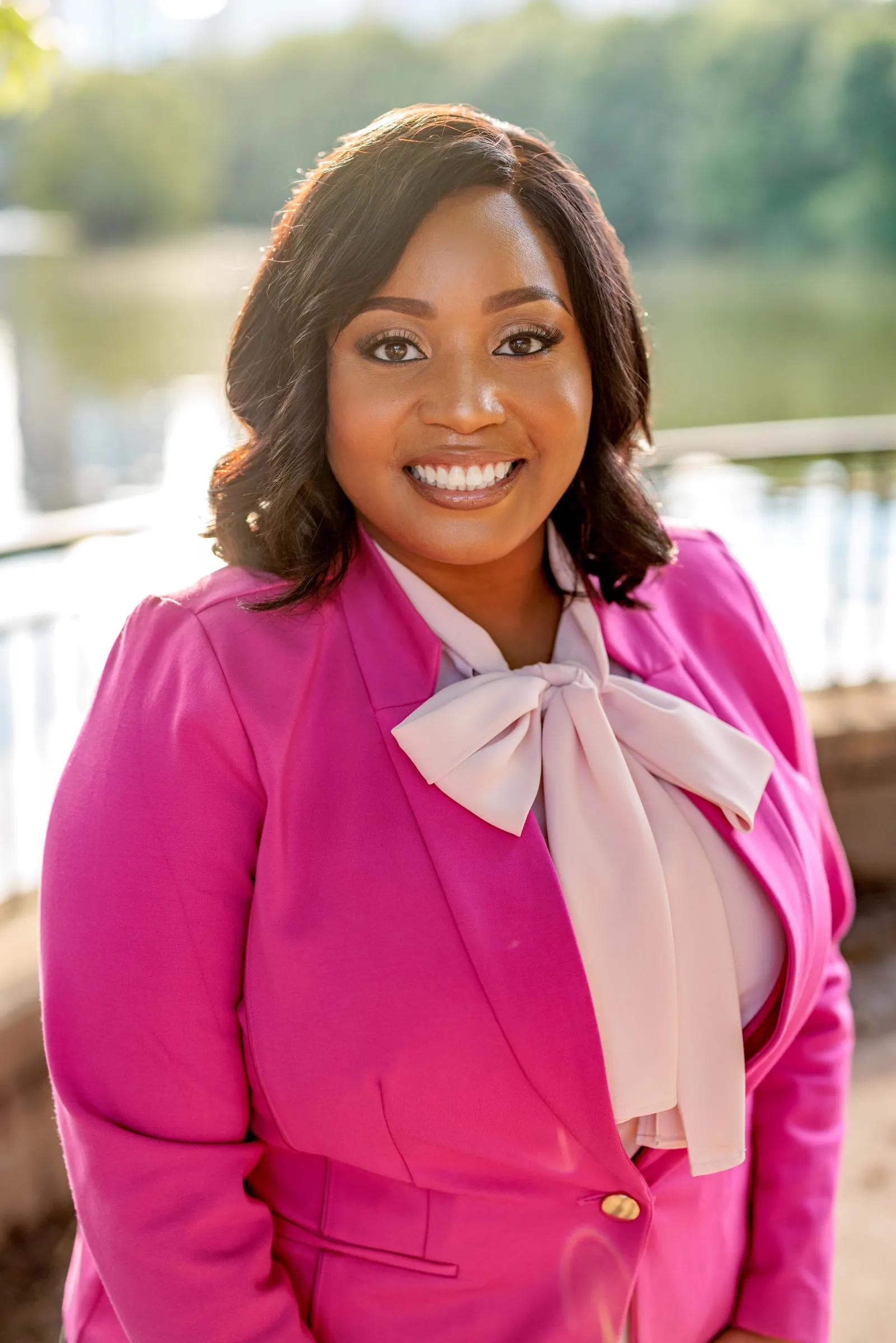 A woman in a bright pink blazer and bow blouse smiling before a summer lake.