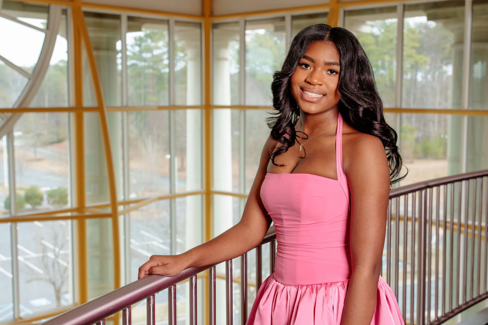 A young woman in a pink dress on a glass-walled stairwell overlooking the trees.