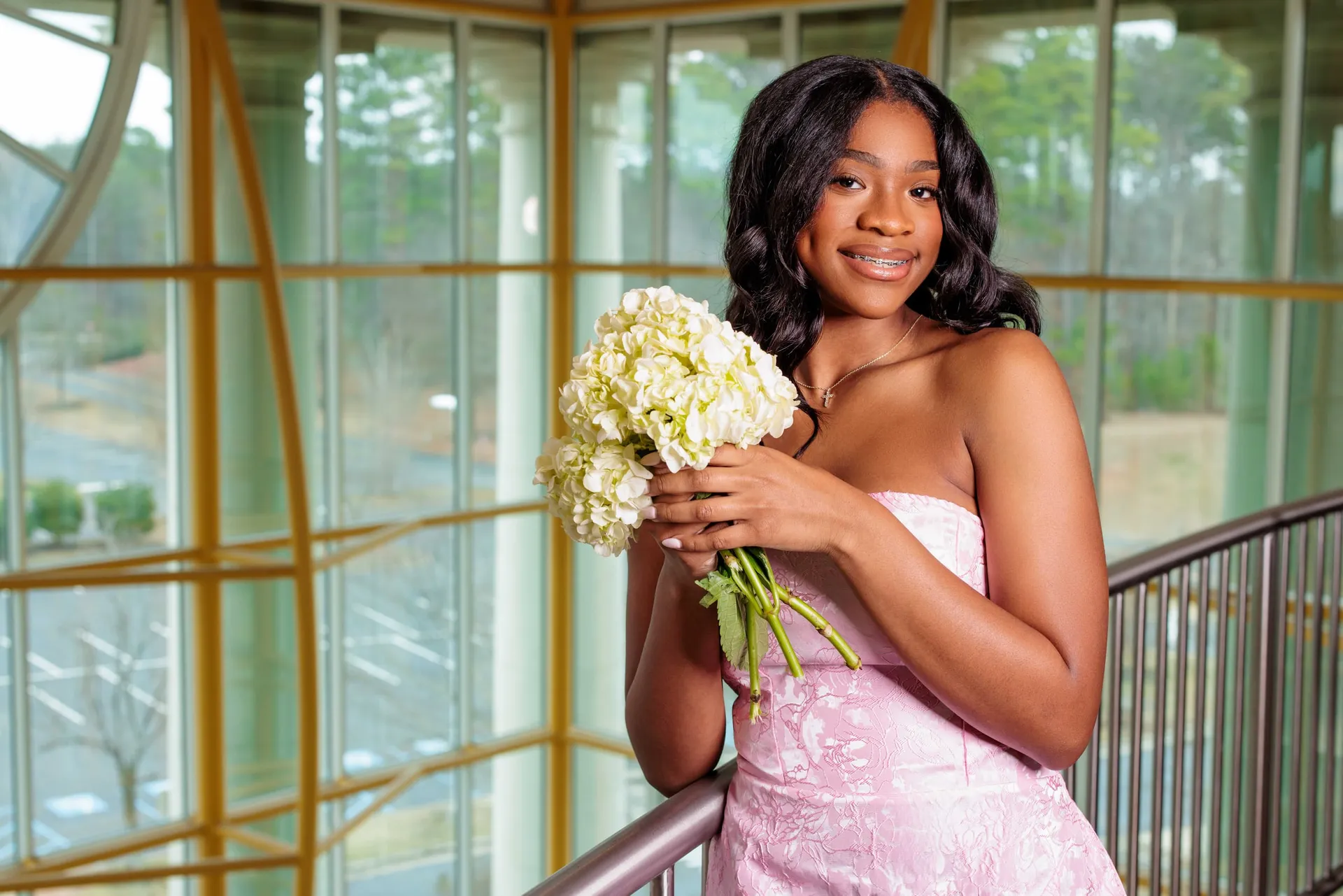 A young woman in pale pink holding white hydrangeas on a glass-walled landing.
