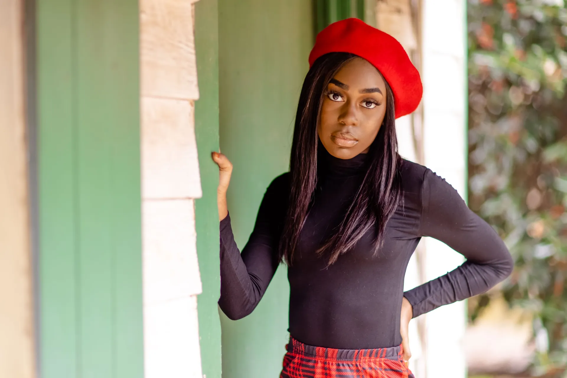 A young woman in a red beret and black turtleneck posing by a green porch column.