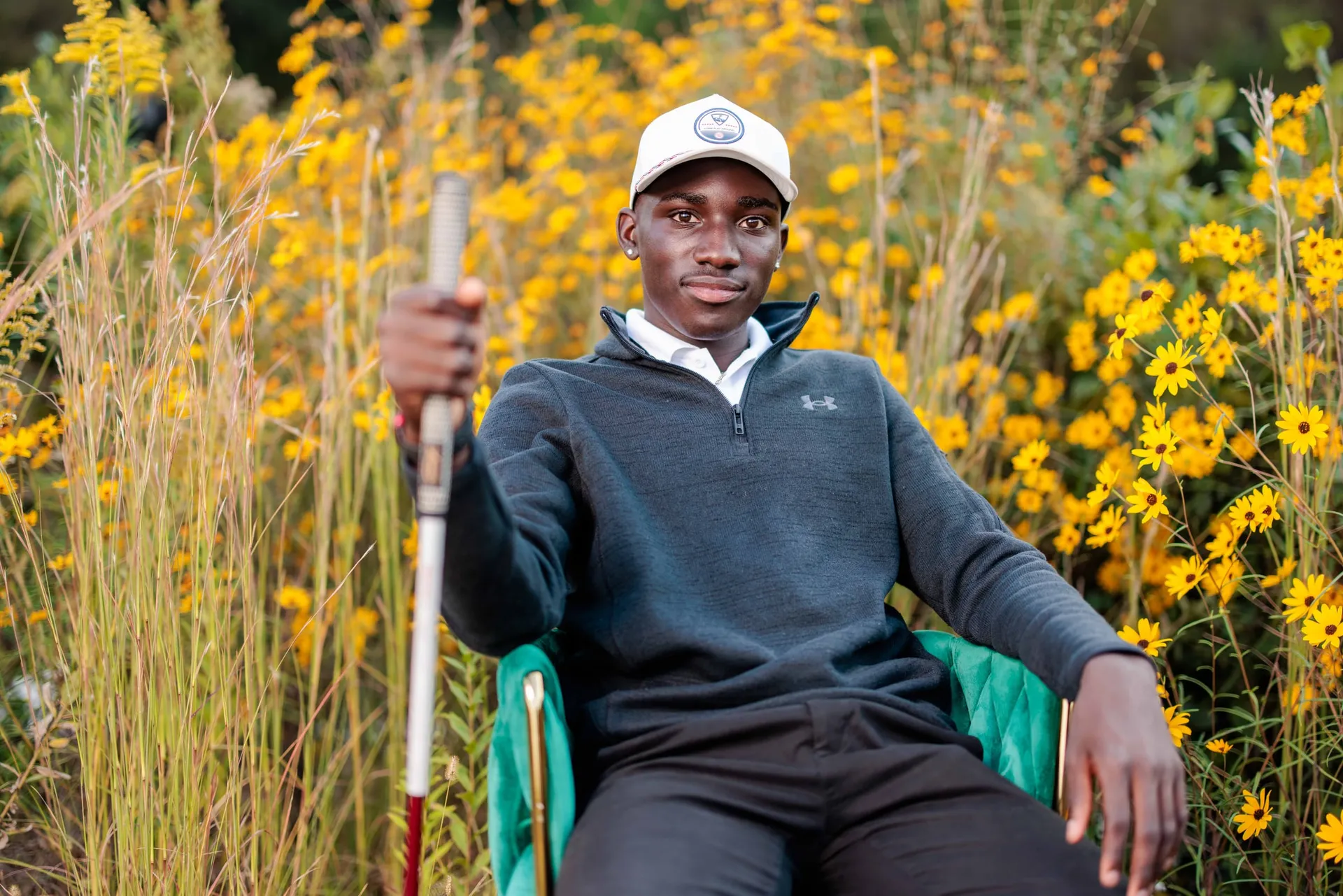 A senior golfer in a white cap seated in a green chair among yellow wildflowers.