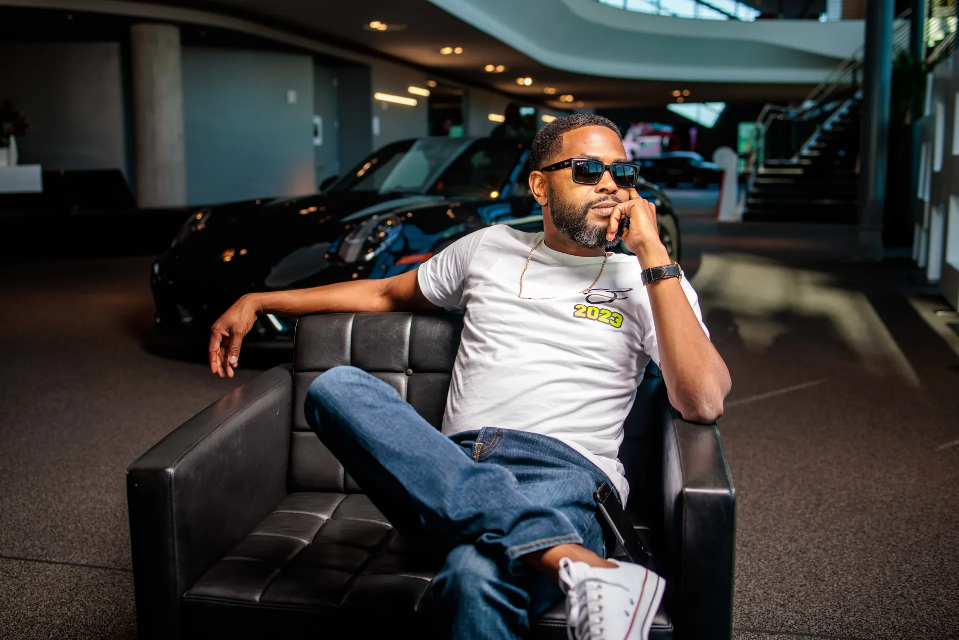 A man in sunglasses and a white tee in a leather chair by a sports car.