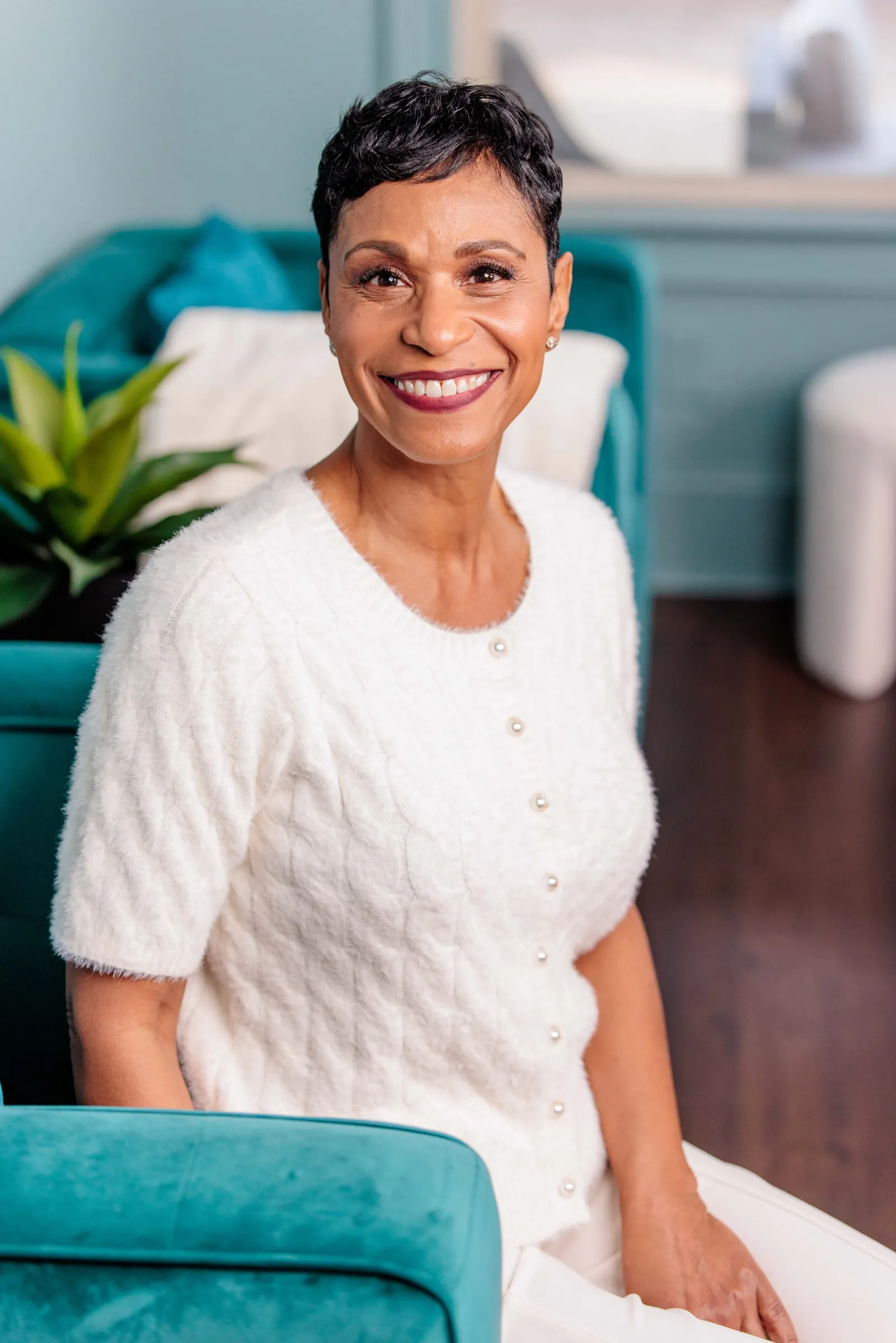 A woman in a white cardigan smiling from the arm of a teal velvet sofa.