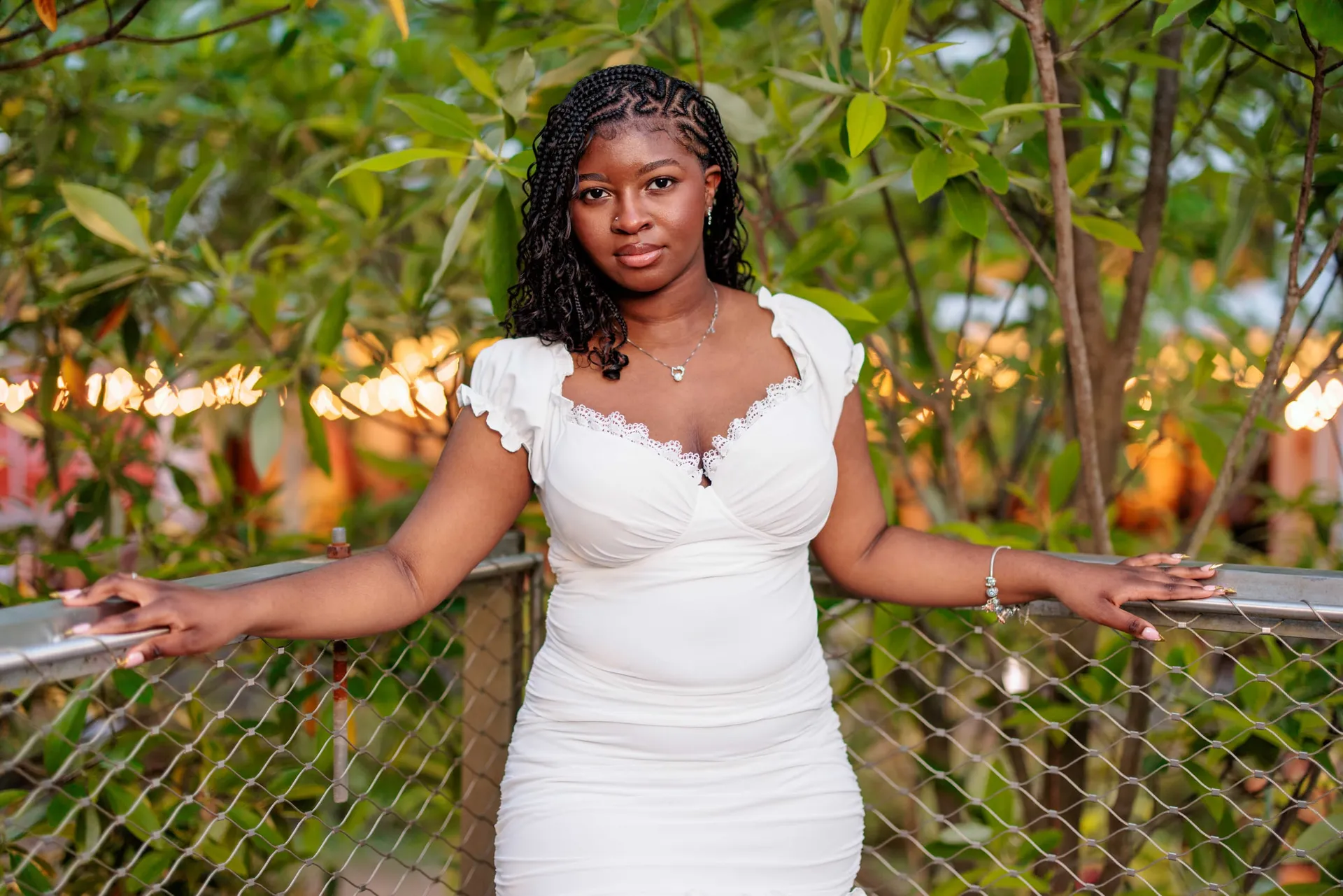 A young woman in a white dress leaning back on a chain-link fence among sunlit greenery.