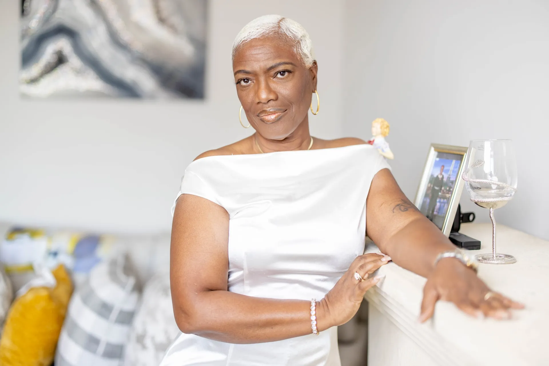 A woman with platinum hair in white satin leaning on a sofa, wine glass beside her.